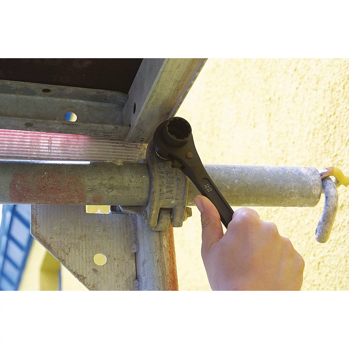 Hand mit schwarzem Werkzeug an einem Gerüst. Metallkonstruktion, gelbe Wand im Hintergrund.